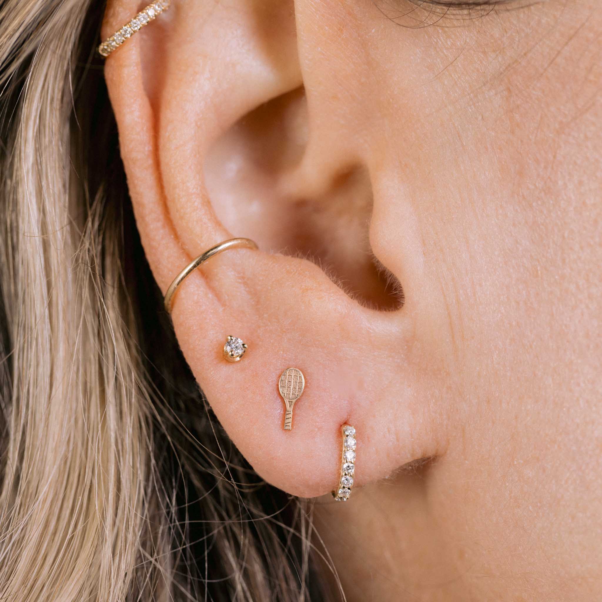 close up of a woman's ear wearing a ZoÃ« Chicco 14k Gold Itty Bitty Tennis Racket Stud Earring in her second piercing layered with a small diamond stud and a Pave Diamond Huggie Hoop