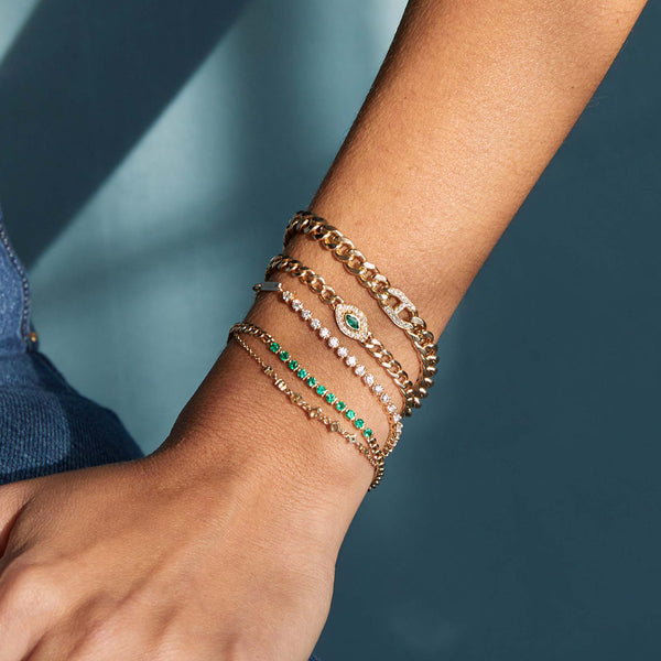 a close up of a woman's arm against a blue background wearing a Zoë Chicco 14k Gold Medium Curb Chain Marquise Emerald Halo Bracelet stacked with four other bracelets on her wrist
