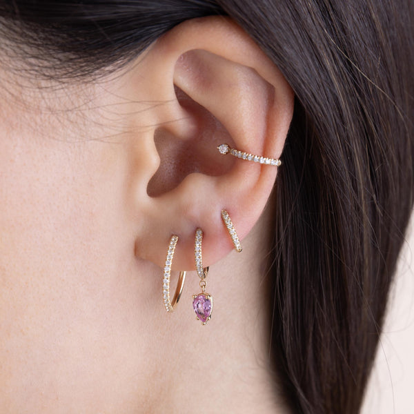 a brunette woman's ear wearing a Zoë Chicco 14k Gold Medium Pavé Diamond Hinge Huggie Hoops with Pear Pink Sapphires in her second piercing with a small and large pavé diamond hoop in her other piercings