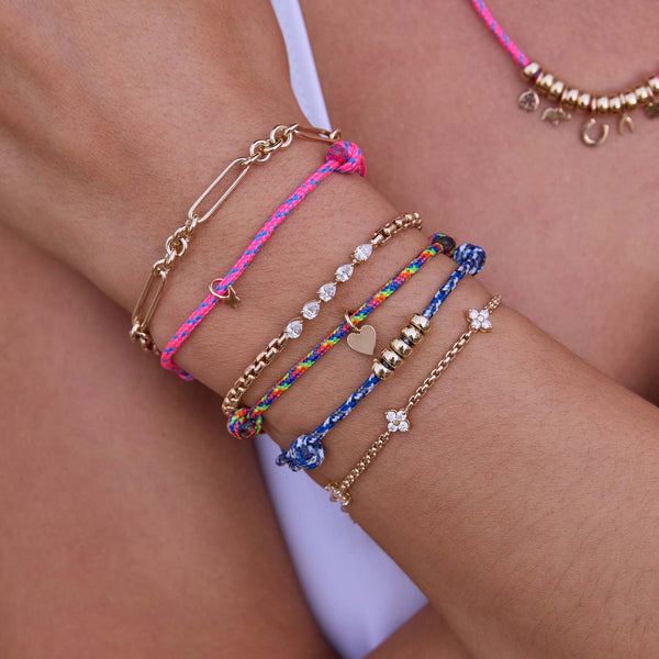 Close-up of a wrist wearing multiple Zoe Chicco colorful paracord bracelets and 14k gold diamond chain bracelets against a neutral background
