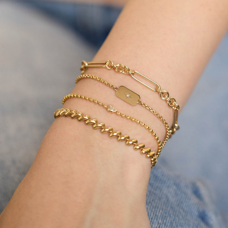 Close up of a person's wrist wearing multiple Zoe Chicco 14k gold chain bracelets on a blurred background