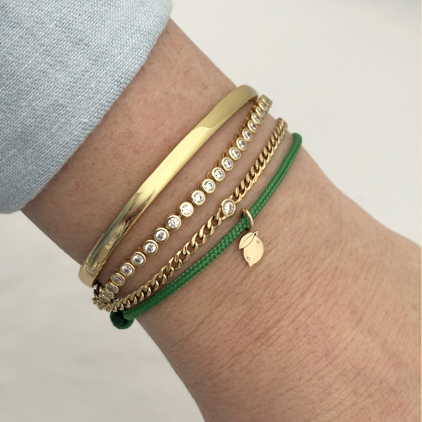 Close-up of a wrist wearing multiple 14k gold and diamond Zoe Chicco bracelets and green paracord bracelet with a Midi Bitty lemon charm on a white background