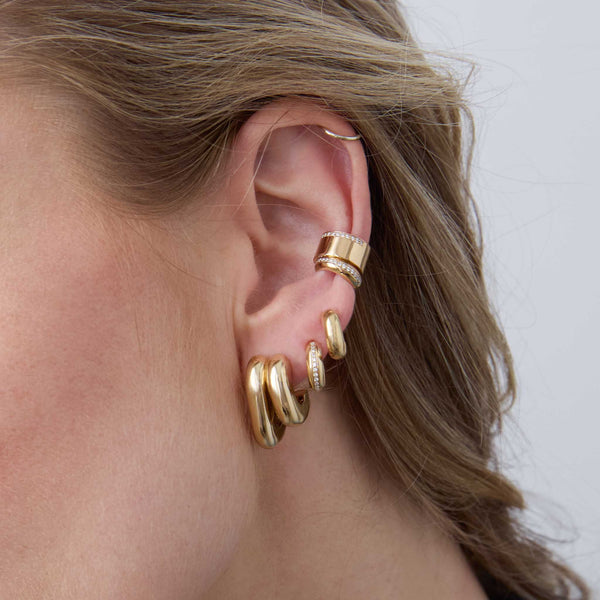 woman's ear wearing a Zoë Chicco 14k Gold Mixed Double Tube Split Oval Hoop Earring layered with a 14k Pavé Diamond Line Chubby Huggie Hoop in her second piercing, 14k Gold Chubby Huggie Hoop in her third, and a 14k Pavé Diamond Line Chubby Ear Cuff and 14k Pavé Diamond Wide Flat Ear Cuff