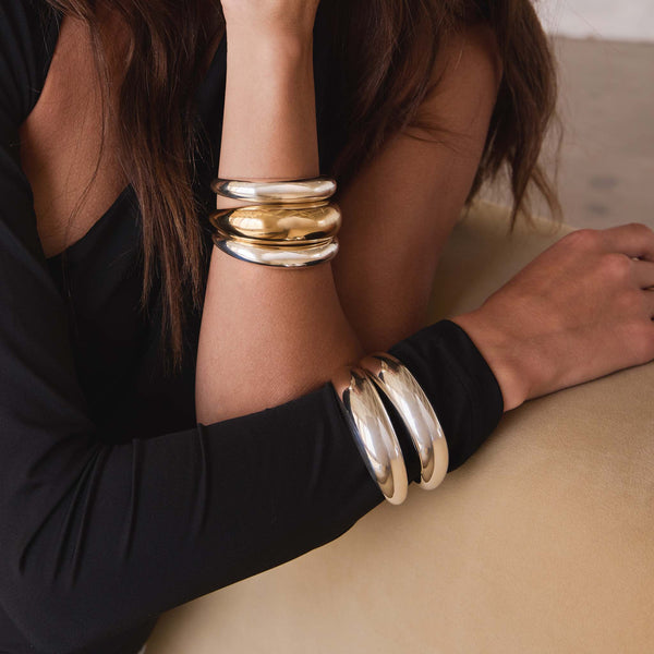 woman in a black dress leaning against a sofa wearing 2 Zoë Chicco Sterling Silver Medium Aura Cuff Bracelets stacked with a 14k Gold Large Aura Cuff Bracelet on one wrist and 2 Sterling Silver Large Aura Cuff Bracelets stacked on her other wrist