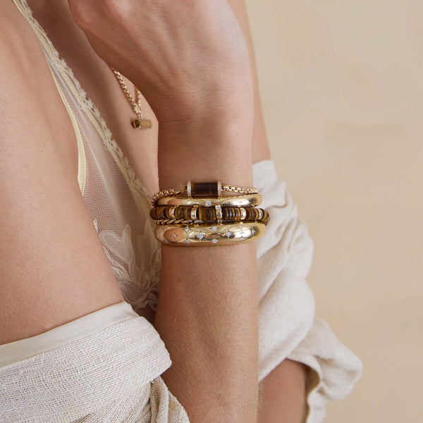 Close-up of a wrist wearing gold bracelets with a neutral background featuring 14k Gold & Tiger Eye Heishi Bead Bracelet with 2 Prong Diamonds
