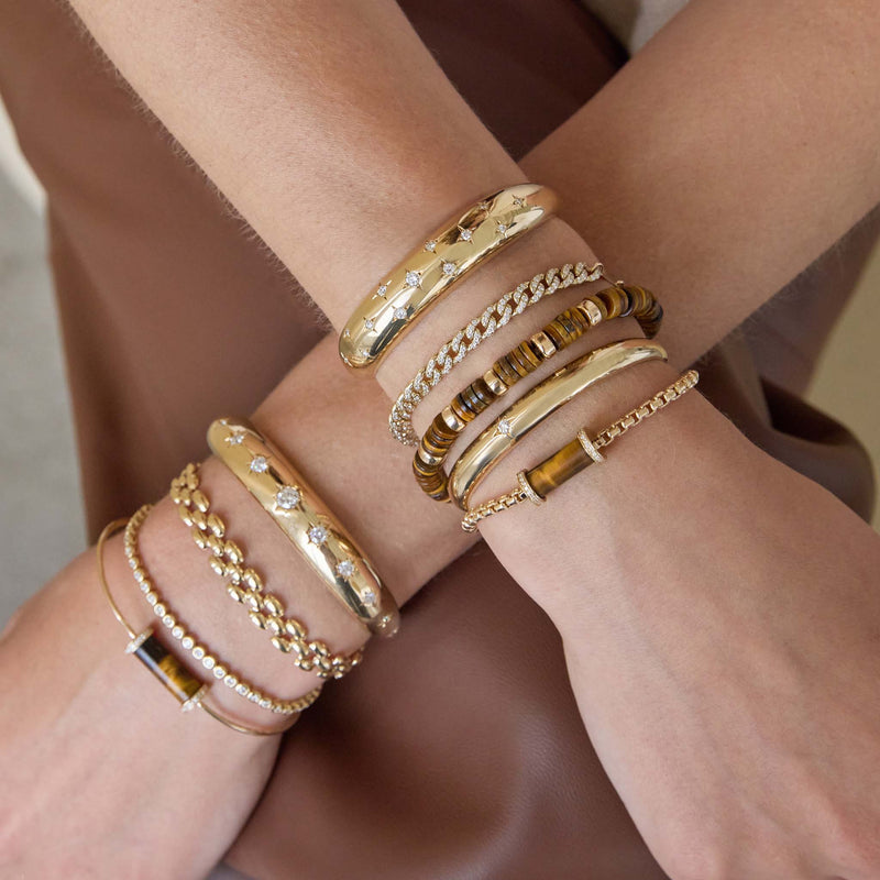 Multiple gold bracelets on a wrist with a neutral background featuring 14k Gold & Tiger Eye Heishi Bead Bracelet