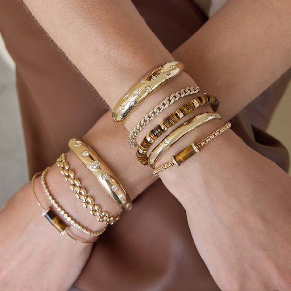 Multiple gold bracelets on a wrist with a neutral background featuring 14k Diamond Tiger Eye Tube Large Box Chain Bracelet