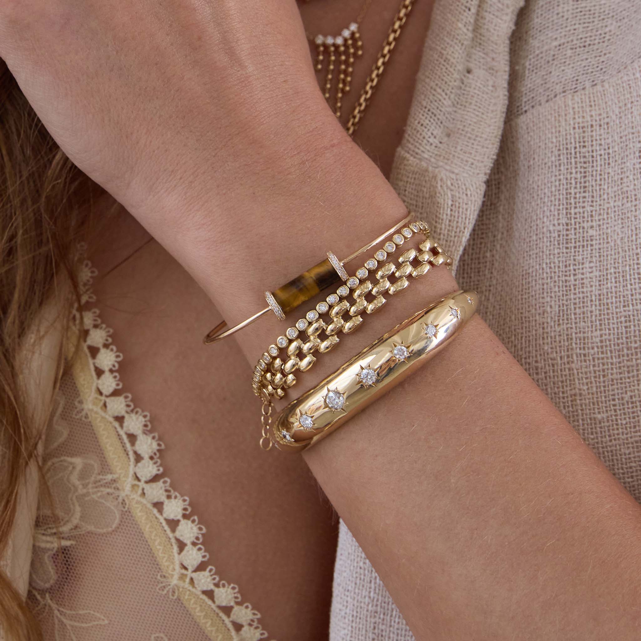 Close-up of a wrist wearing 14k Diamond Tiger Eye Tube Cuff and gold bracelets with a neutral background