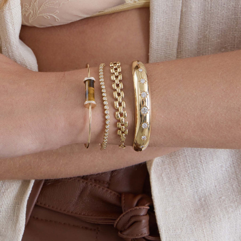 Close-up of a wrist wearing 14k Diamond Tiger Eye Tube Cuff and gold bracelets with a neutral background