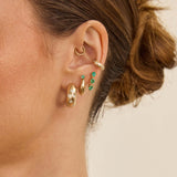 Close-up of an ear wearing multiple Zoe Chicco 14k gold hoop earrings with diamonds and emeralds on a neutral background