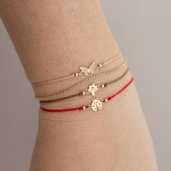 A woman is wearing 14k Midi Bitty Butterfly, 14k Midi Bitty Mushroom Cord Bracelet , 14k Midi Bitty Ladybug Cord Bracelet