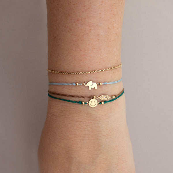 A woman wears 14k Midi Bitty Elephant Cord Bracelet, 14k Midi Bitty Eye Cord Bracelet and 14k Midi Bitty Smiley Face Cord Bracelet