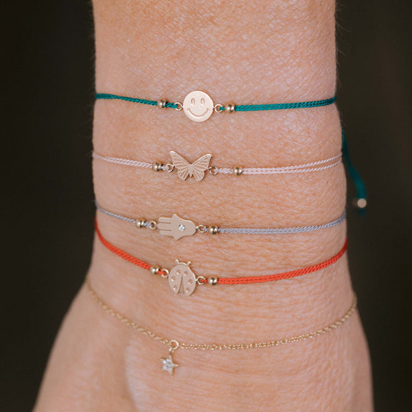 a close up of a woman's wrist against a black background wearing a Zoë Chicco 14k Gold Midi Bitty Smiley Face Cord Bracelet layered with a Zoë Chicco 14k Gold Midi Bitty Butterfly, Diamond Hamsa, and Ladybug Cord Bracelets