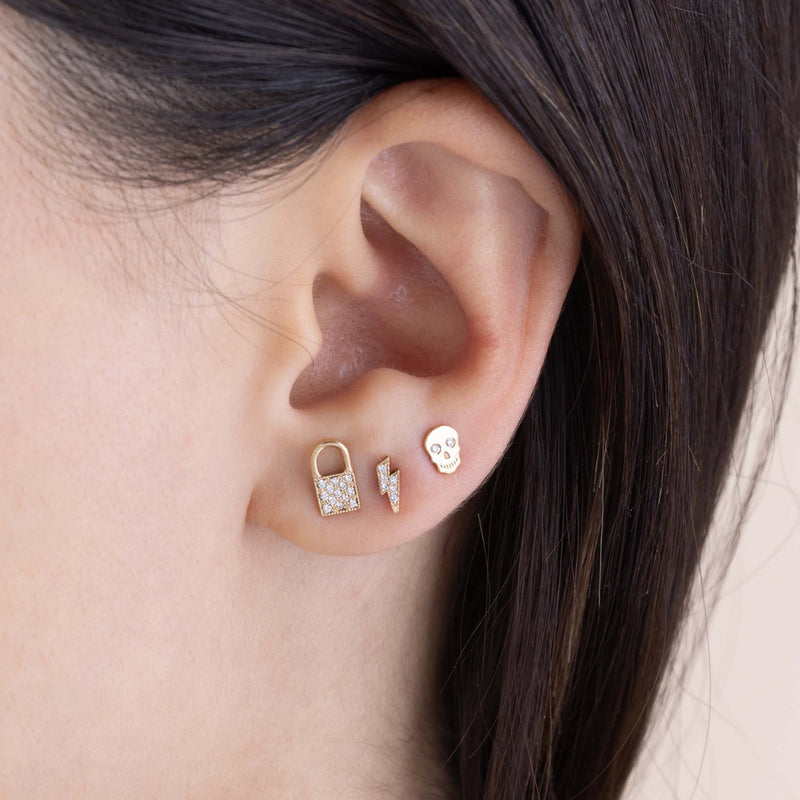 A woman with dark hair is wearing 14k Midi Bitty Pavé Diamond Padlock Stud, 14k Itty Bitty Pavé Diamond Lightning Bolt Stud and 14k Itty Bitty Skull with Diamond Eyes Stud