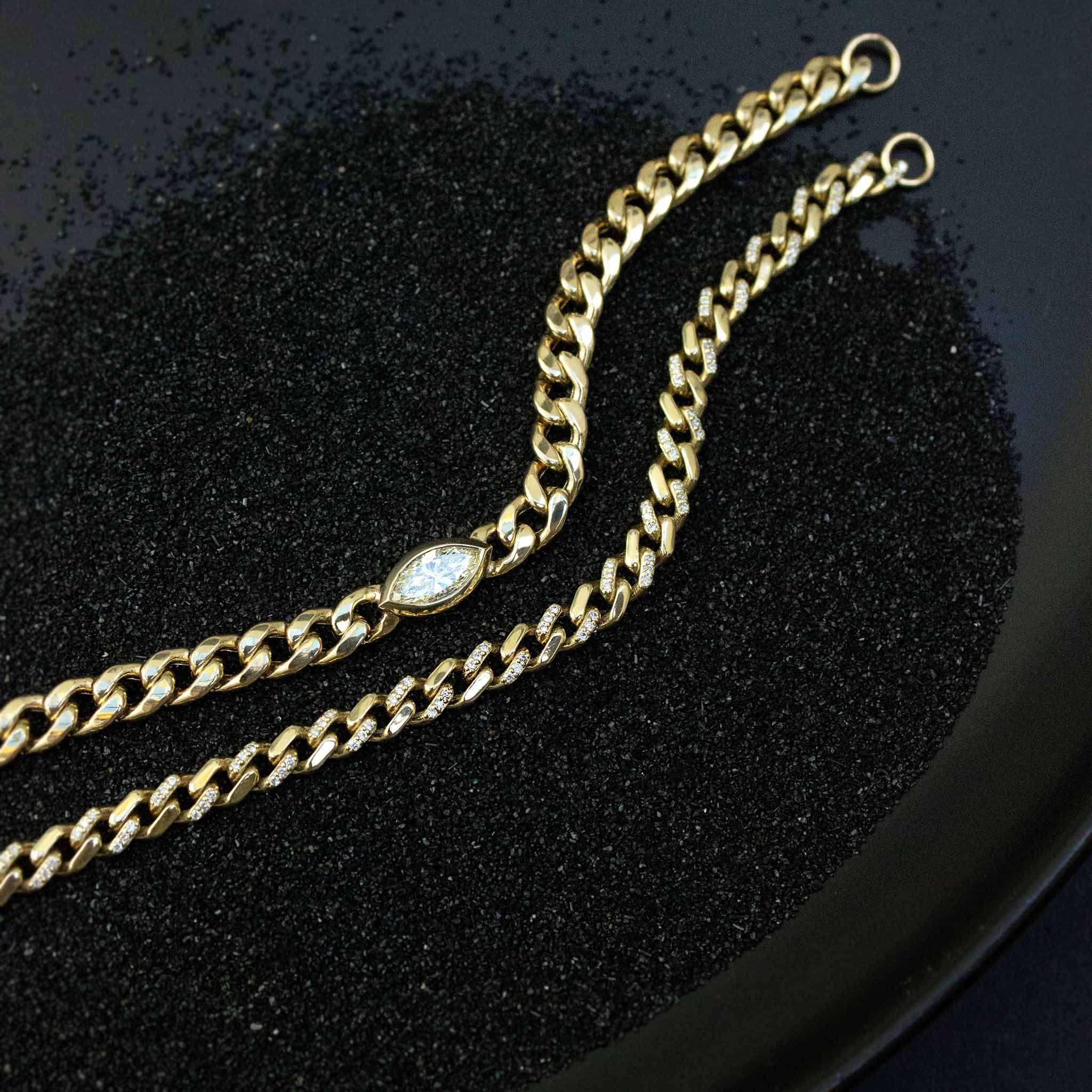 On a black background of sand lies two curb chain bracelets: One of a Kind Marquise diamond bracelet centered on a large curb chain and a medium curb chain with alternating pave diamond links.  