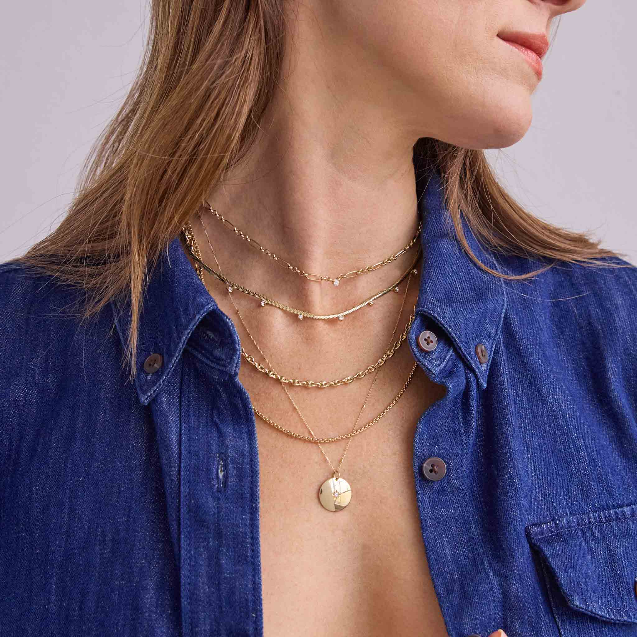 Woman wearing multiple layers of Zoe Chicco 14k gold necklaces with diamonds and wearing a blue denim shirt against a light gray background