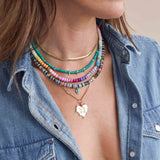 woman in a denim shirt wearing a Zoë Chicco 14k Gold Mixed Dark Gemstone Rondelle Bead Necklace with Pear Turquoise Pendant layered with a Medium Snake Chain, 3 Prong Diamond Turquoise Bead Necklace, a Rainbow Opal Rondelle Bead Necklace, and a 14k Gold and Prong Diamond Bar Necklace with a 7 Prong Diamond Heart with Pavé Diamond Love Pendant clipped onto the necklace