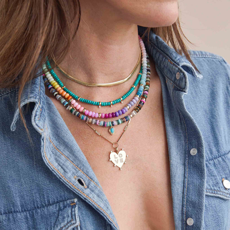 woman in a denim shirt wearing a Zoë Chicco 14k Gold Mixed Dark Gemstone Rondelle Bead Necklace with Pear Turquoise Pendant layered with a Medium Snake Chain, 3 Prong Diamond Turquoise Bead Necklace, a Rainbow Opal Rondelle Bead Necklace, and a 14k Gold and Prong Diamond Bar Necklace with a 7 Prong Diamond Heart with Pavé Diamond Love Pendant clipped onto the necklace
