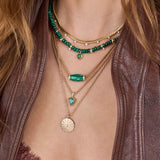 Close-up of layered necklaces 14k Diamond Malachite Barrel Bead Medium Box Chain Necklace, emerald and malachite necklaces, and gold pendants on a person wearing a brown leather jacket.