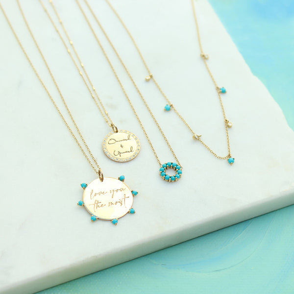 a Zoë Chicco 14k Gold Medium "love you the most" Disc with Prong Turquoise Charm Pendant hanging off a cable chain necklace laying flat on a marble tray