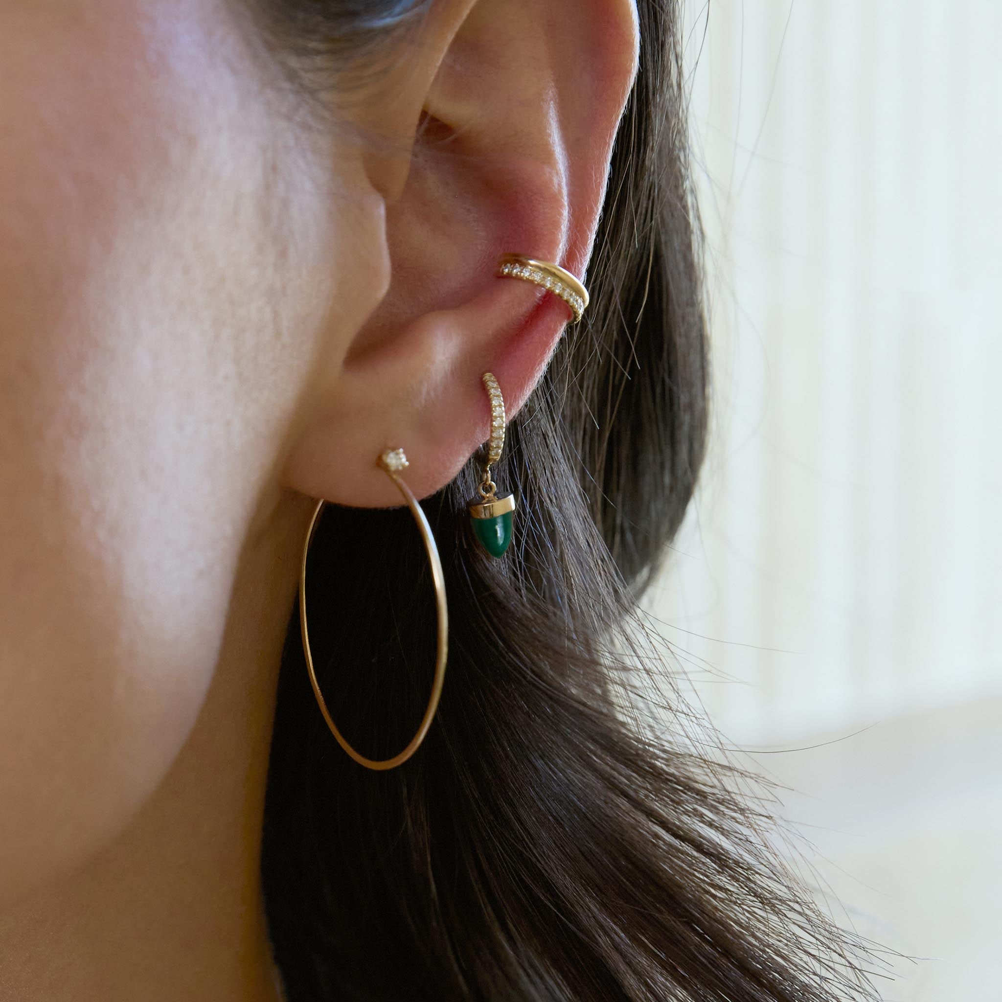 Close-up of an ear wearing gold hoop earrings with small white diamonds and a malachite gemstone bullet with an ear cuff