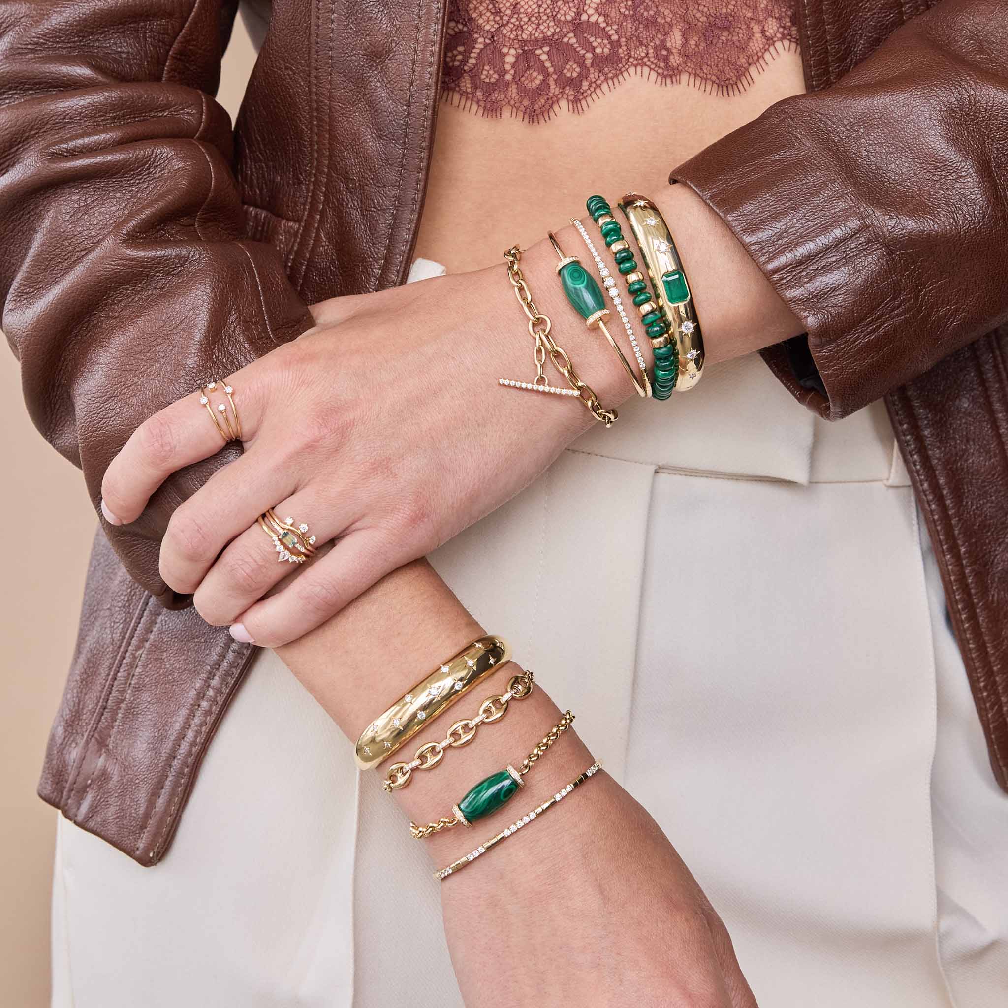 Close-up of a person's arm wearing multiple bracelets and rings with a brown leather jacket and lace top featuring 14k Diamond Malachite Barrel Bead Cuff