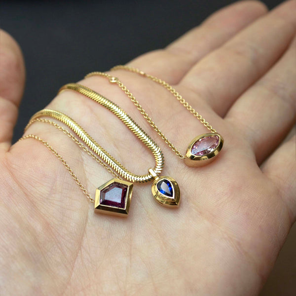 a woman's palm face up with a Zoë Chicco 14k Gold Pear Blue Sapphire Bezel Pendant Snake Chain Necklace laying in her palm next to 2 one of a kind pink sapphire gemstone necklaces
