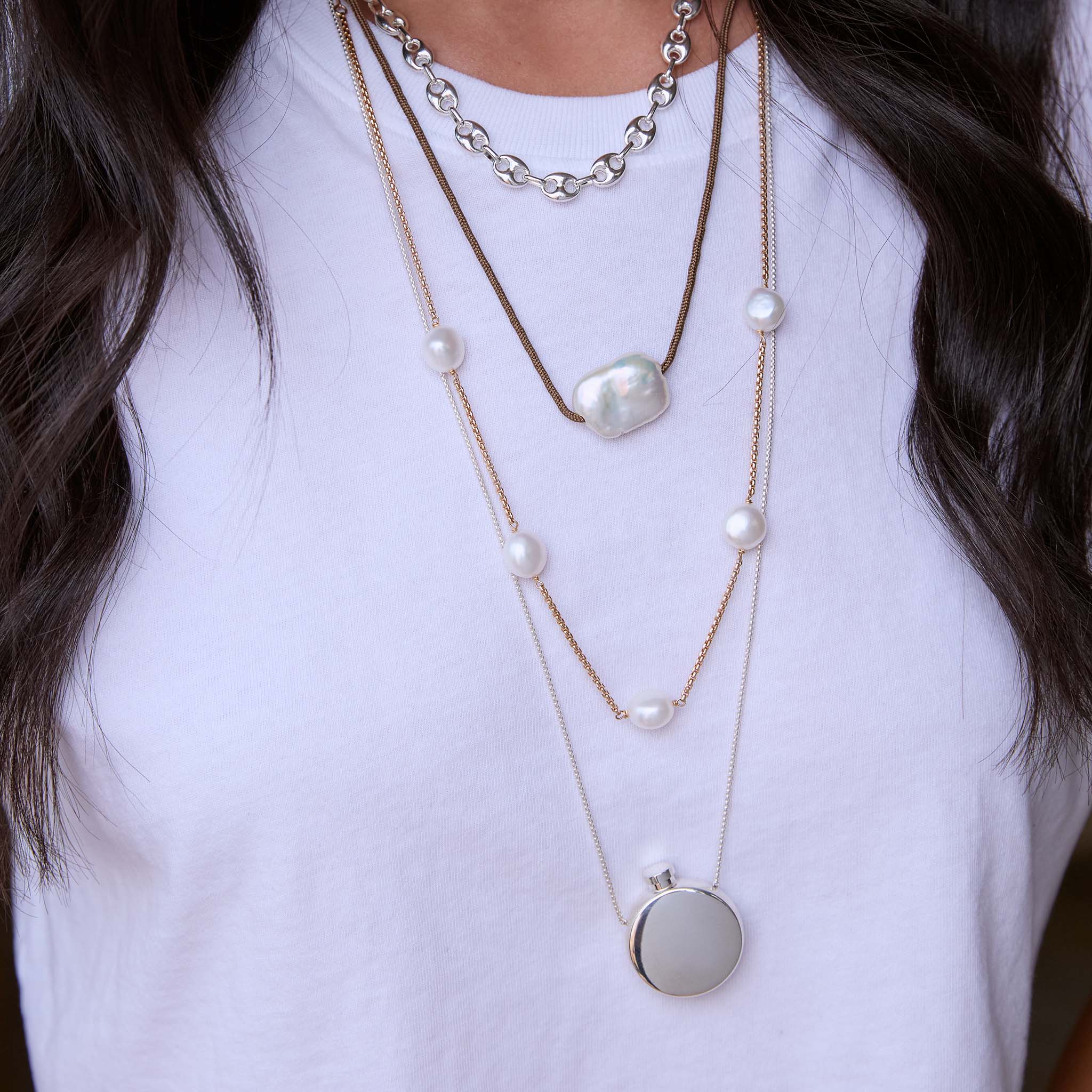 Close up of a neck wearing multiple Zoe Chicco layered sterling silver and gold chain necklaces with pearls against a white t-shirt