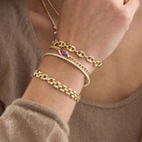 Close up of a wrist wearing multiple Zoe Chicco 14k gold pink sapphire bracelets and diamond tennis flex bracelet on a neutral background