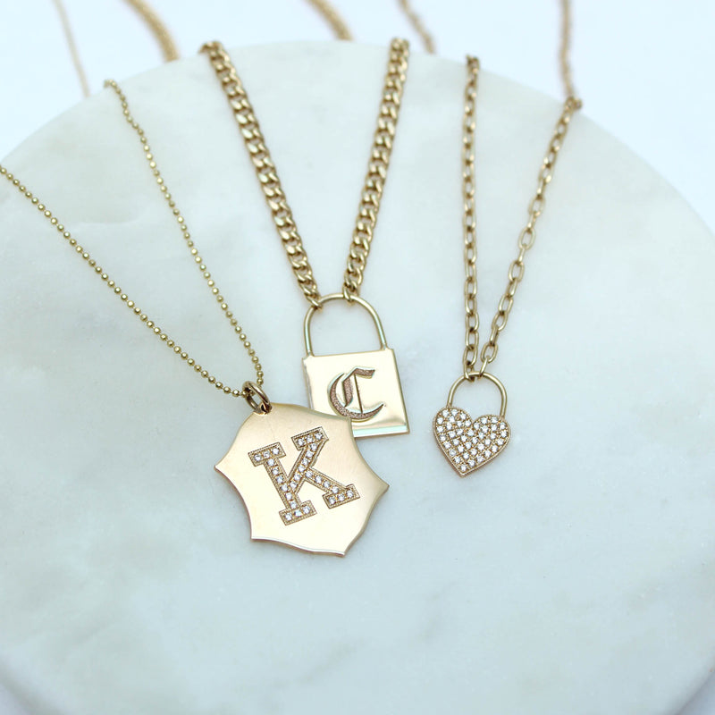 a Zoë Chicco 14kt Gold Large Engraved Initial Padlock Small Curb Chain Necklace engraved with the letter C laying flat on a marble tray with two other necklaces