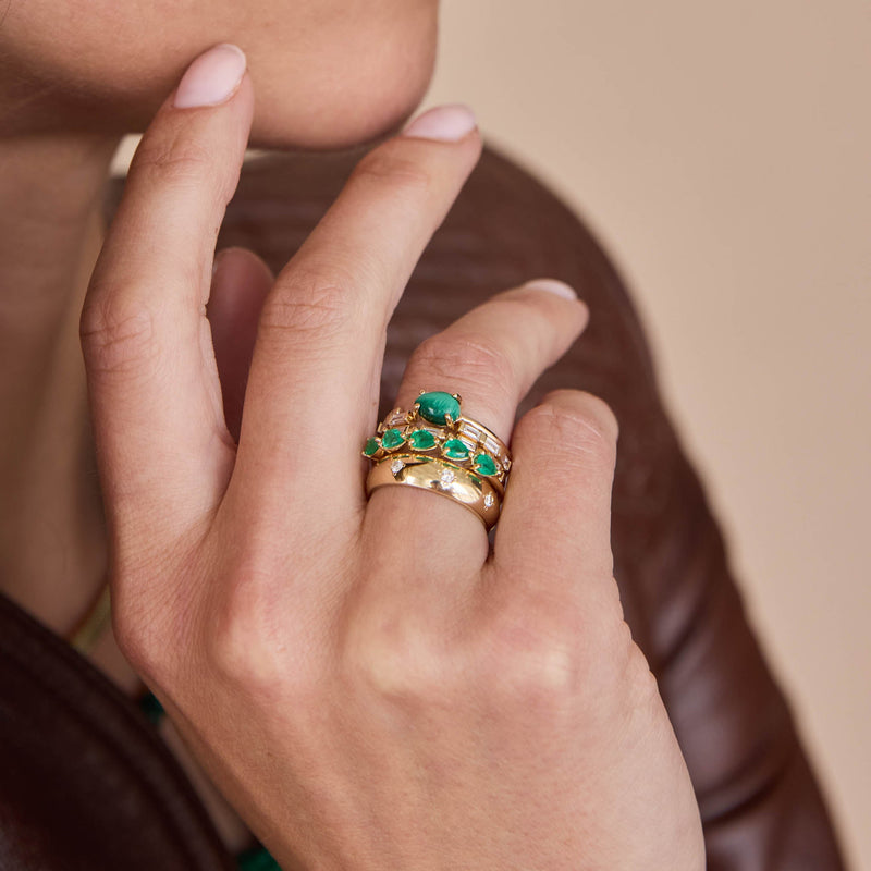 Close up of a hand wearing Zoe Chicco 14k gold rings with diamonds and pear-shaped green emeralds on a neutral background
