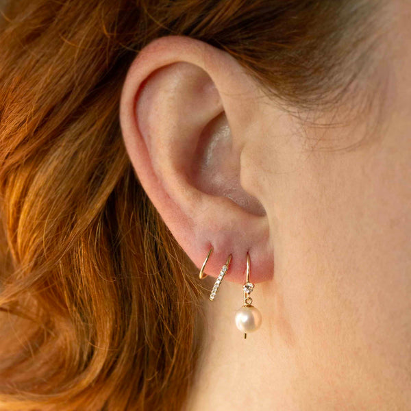 a close up of a woman's ear wearing a Zoë Chicco 14k Gold Prong Diamond & Dangling Pearl Hook Earring layered with two hoops