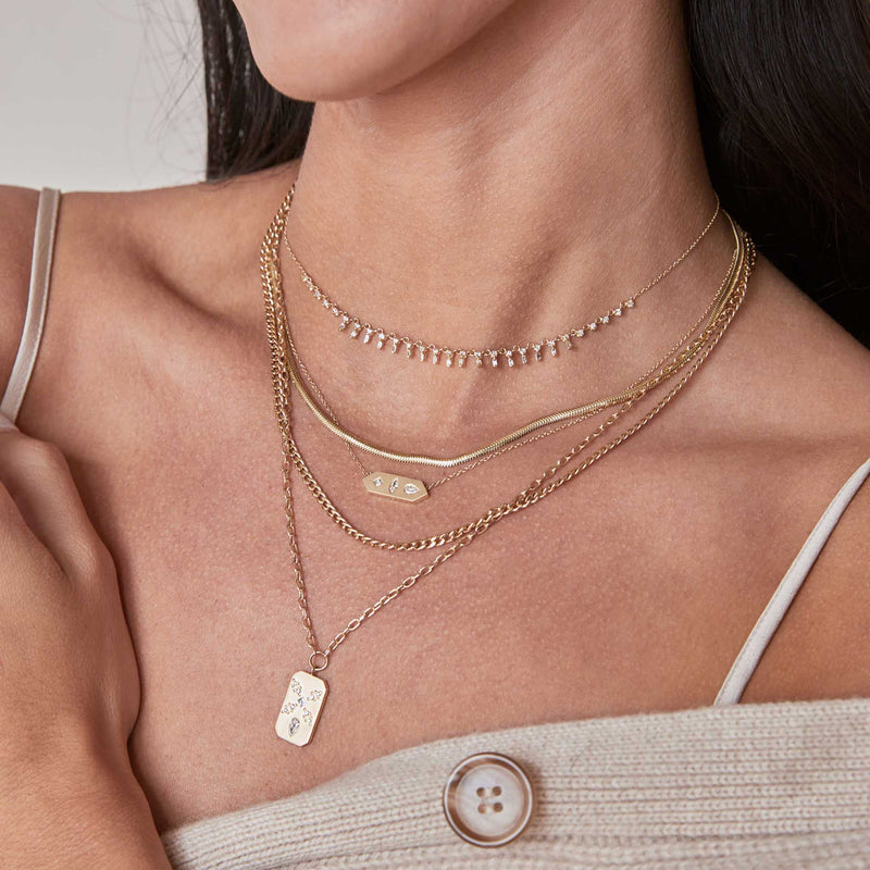 woman in a beige sweater wearing a Zoë Chicco 14k Gold Diamond Mosaic Brushed Gold Horizontal Hexagon Tag Necklace layered with four other necklaces