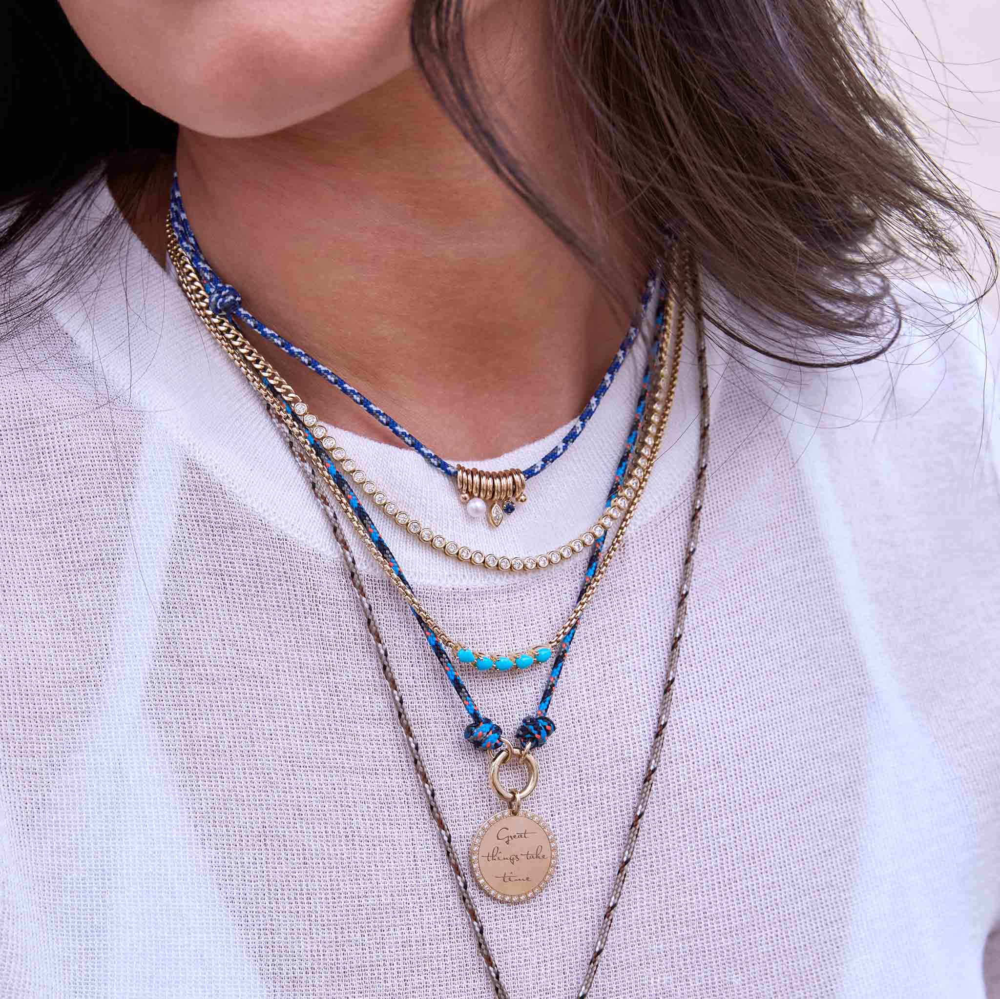 Person wearing multiple Zoe Chicco 14k gold and diamond necklaces and blue shades of paracord necklaces against a white textured shirt