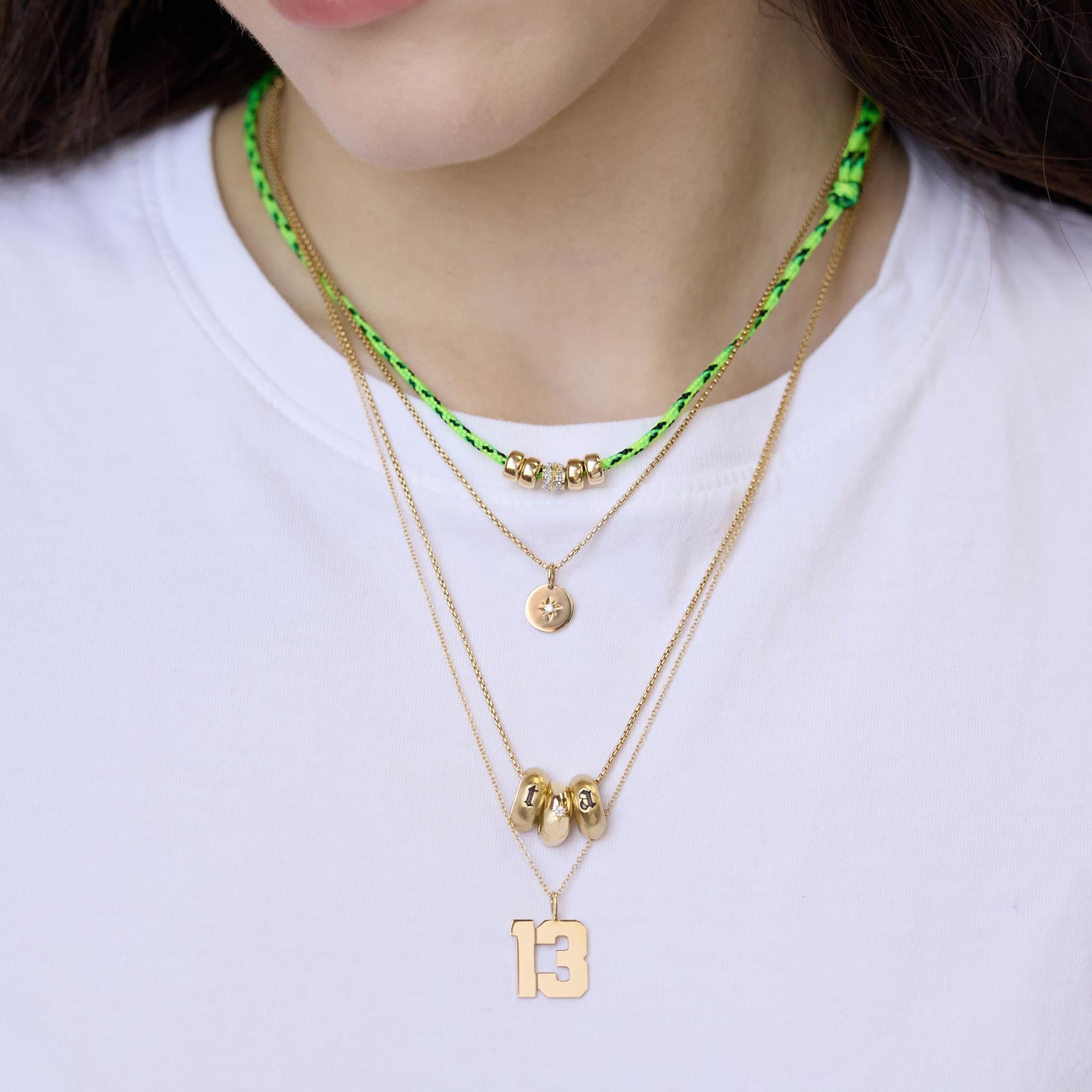 woman in a white tshirt wearing a 14k 2 Initial Letter & Star Set Diamond Half Round Slide Bead Box Chain Necklace with the letter "t" and "a" layered with a 14k 6mm Gold & 1 Double Row Diamond Rondelle Bead Paracord Necklace, 14k Star Set Diamond Disc Box Chain Necklace, and a 14k Gold Custom Large Varsity Number "13" Pendant Necklace

