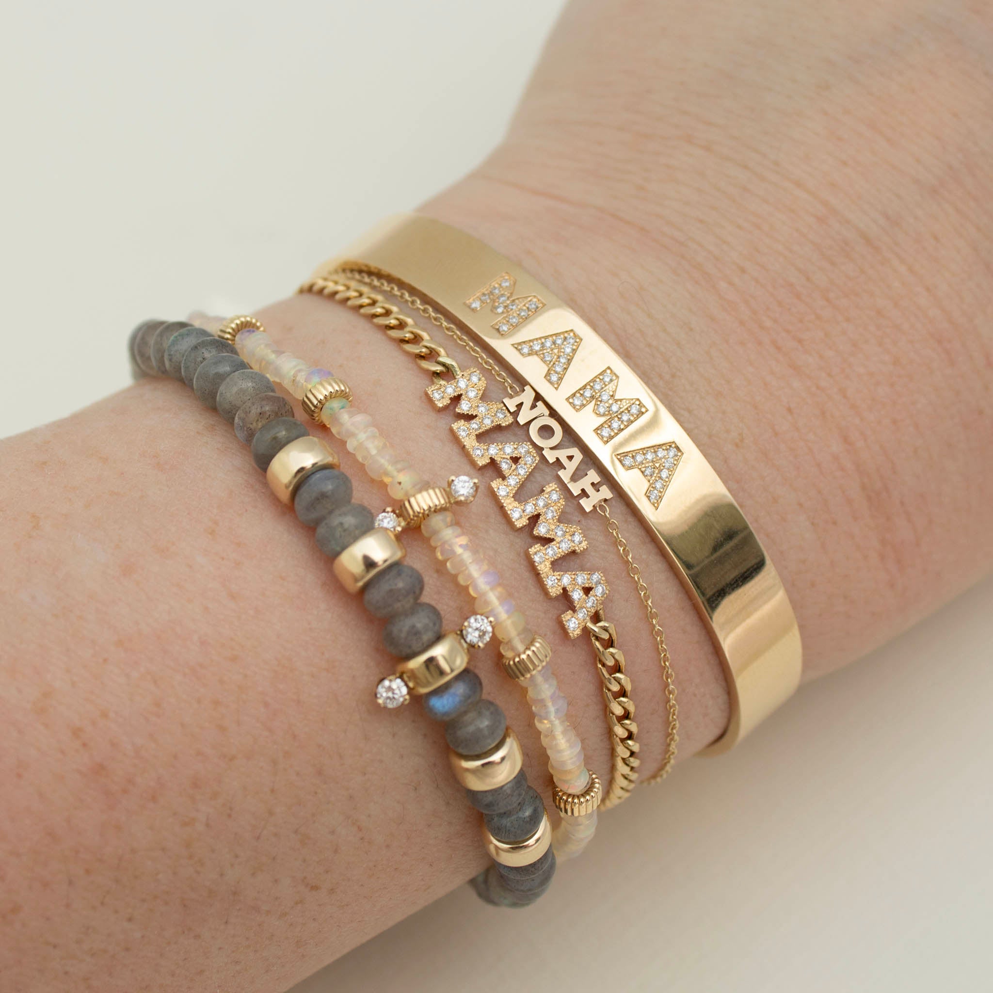 close up of a woman's wrist wearing a Zoë Chicco 14k Gold Personalized Diamond Letters Wide Cuff Bracelet customized with "MAMA" and a Zoë Chicco 14k Gold Custom Itty Bitty Letters Bracelet with the name NOAH layered with a 14k Pavé Diamond MAMA Small Curb Chain Bracelet, 14k Gold & Fire Opal Rondelle Bead Bracelet with 2 Prong Diamonds, and a 14k Gold & Labradorite Rondelle Bead Bracelet with 2 Prong Diamonds