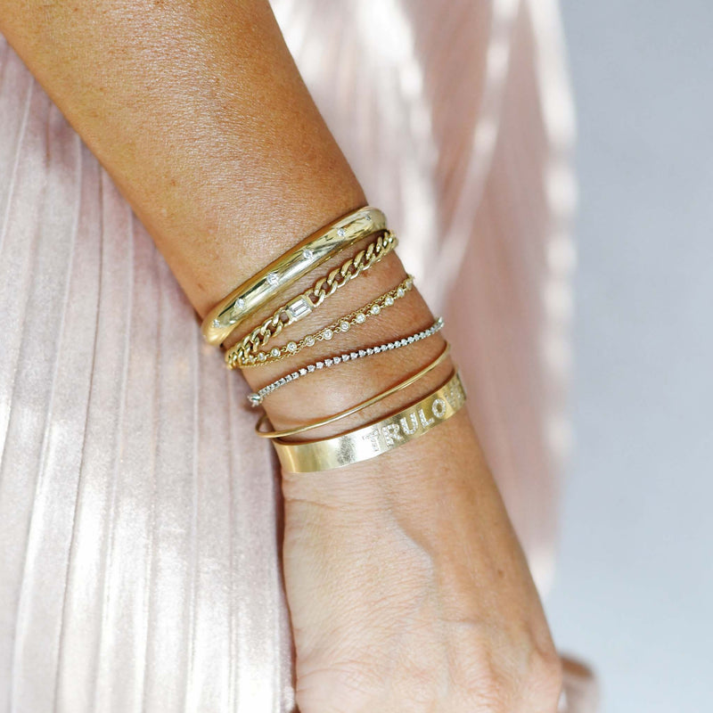 woman in a pink pleated skirt wearing a Zoë Chicco 14k Gold Personalized Diamond Letters Wide Cuff Bracelet customized with "TRULOVE" layered with other gold and diamond bracelets