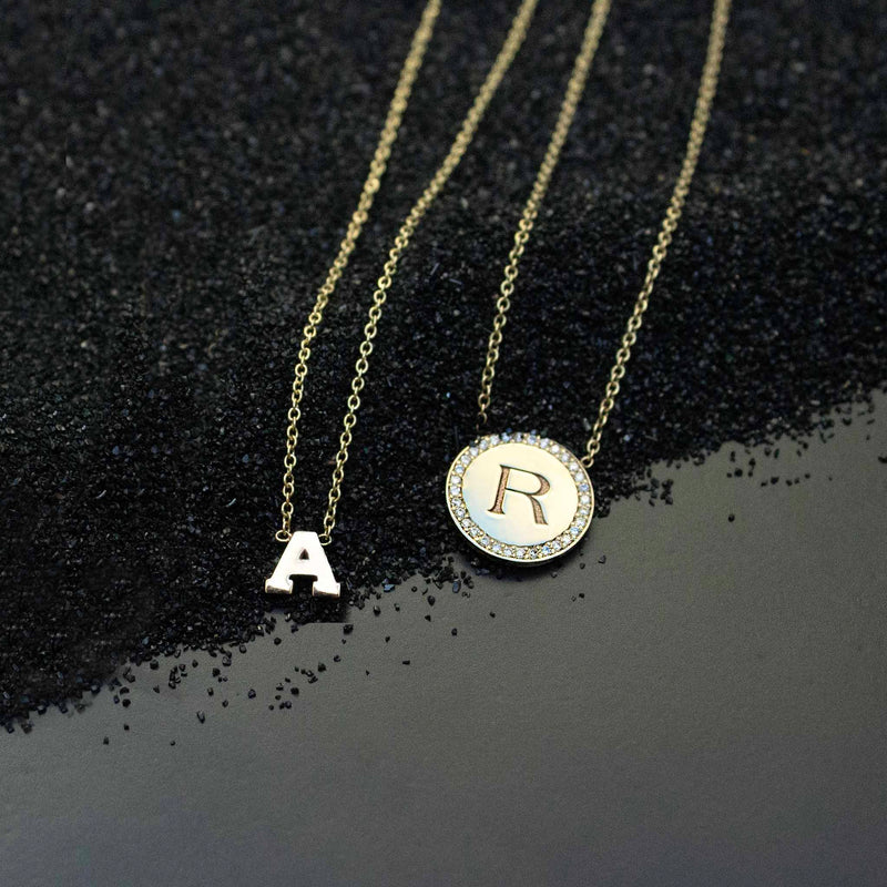 Laying on black sand are two necklaces including a simple block letter necklace and an engraved letter necklace with pave diamond border.