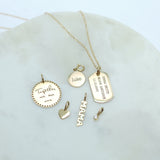 a Zoë Chicco 14k Gold Vertical Text Small Dog Tag Charm on a cable chain laying flat on a marble tray with several other charms