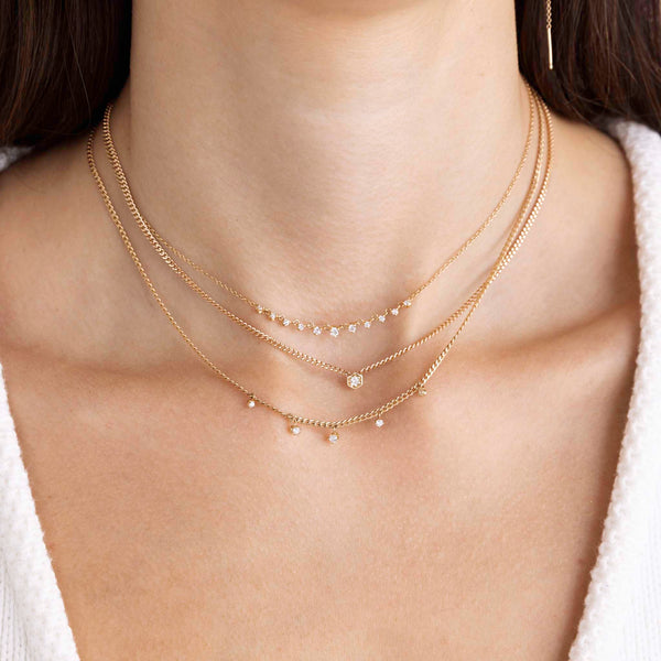 A woman with dark hair is wearing a Zoe Chicco necklace stack including 14k 9 Linked Prong Diamond Necklace, 14k Gold Diamond Hexagon Curb Chain Necklace, and 14k 5 Dangling Prong Diamond Extra Small Curb Chain Necklace