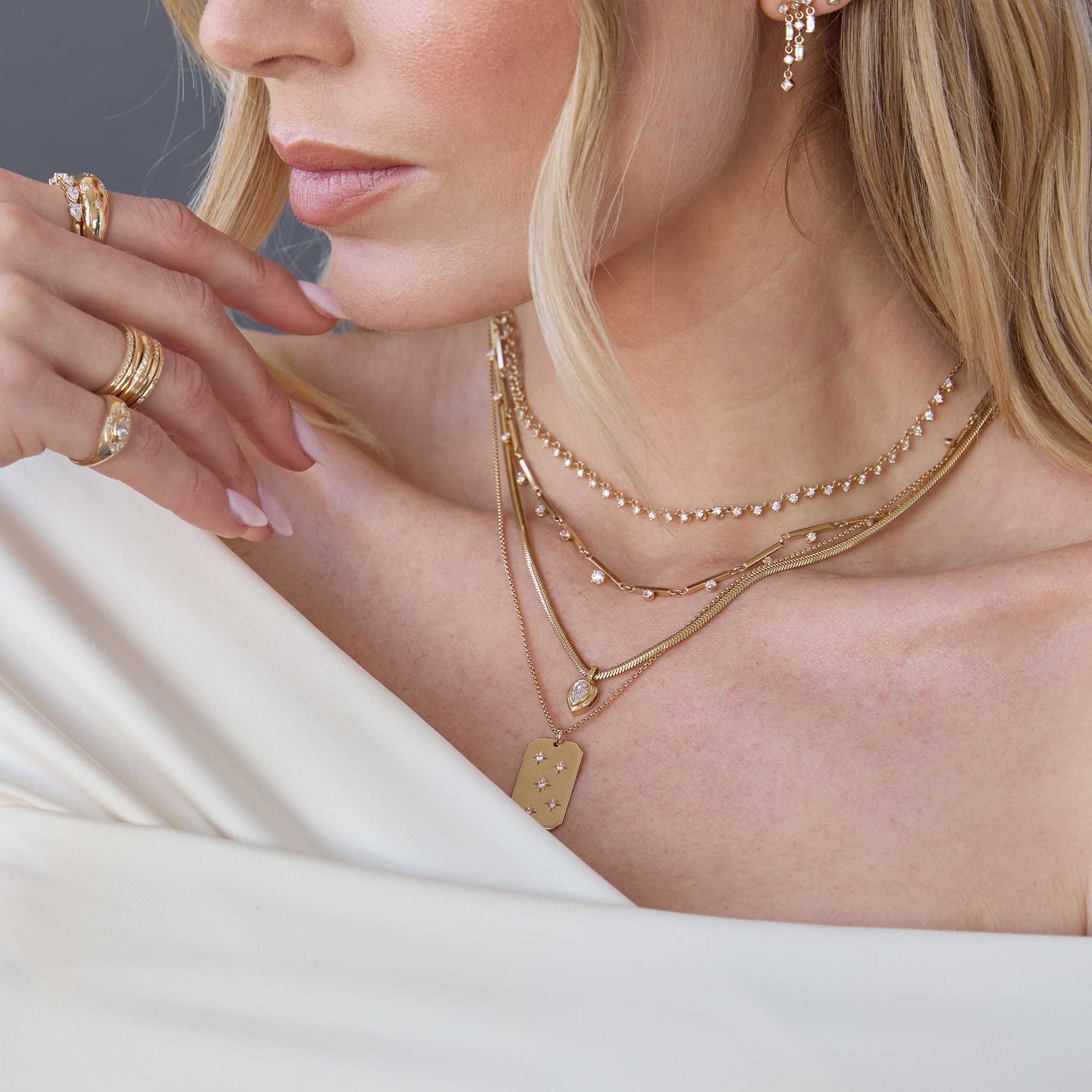 woman in an off shoulder top wearing a Zoë Chicco 14k Gold 15 Graduated Prong Diamond Linked Bar Necklace layered with a Diamond Tennis Necklace, 14k Pear Diamond Bezel Pendant Snake Chain Necklace, and a Large Scattered Star Set Diamond Dog Tag Pendant on a 14k Gold Extra Small Box Chain Necklace
