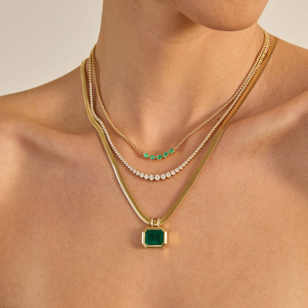 Close-up of a person wearing multiple Zoe Chicco 14k gold necklaces with green emeralds and diamonds on a neutral background
