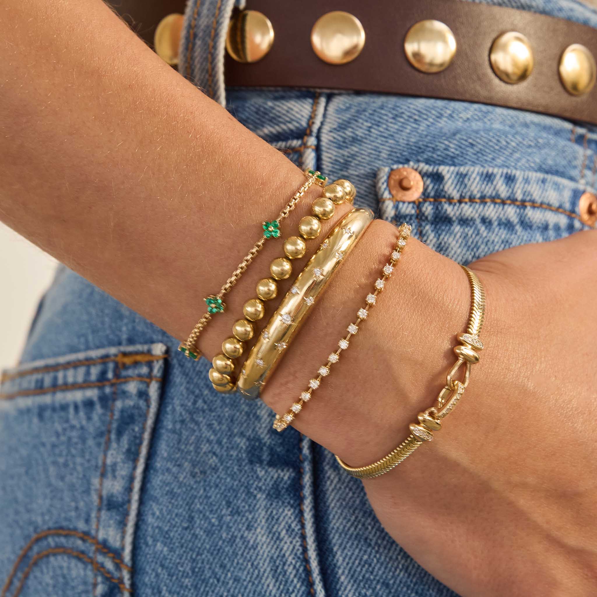 Gold bracelets on a wrist adorn with diamonds and emeralds are shown against a denim background
