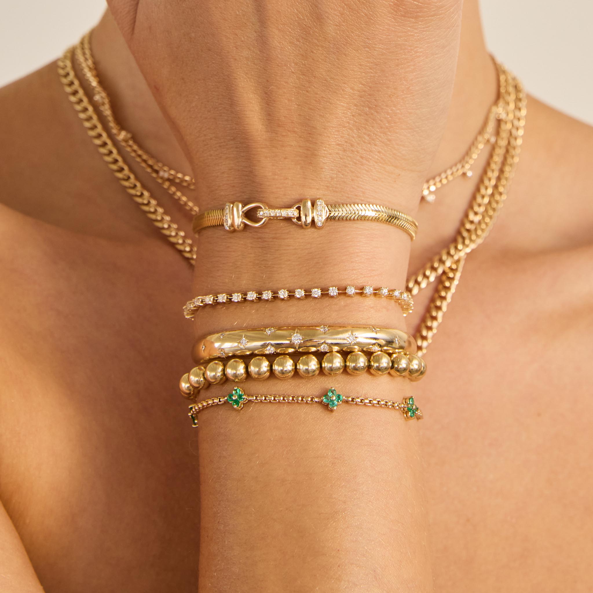 Gold bracelets stacked on a wrist with  diamond and emeralds are shown against a neutral background.
