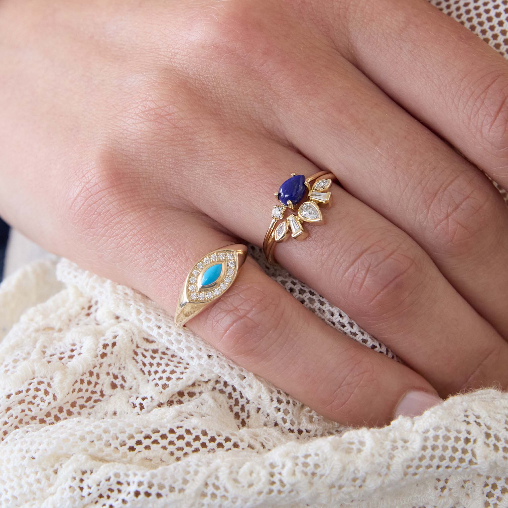 Close up of a hand wearing multiple Zoe Chicco 14k gold and diamond rings with blue gemstones on a white lace background