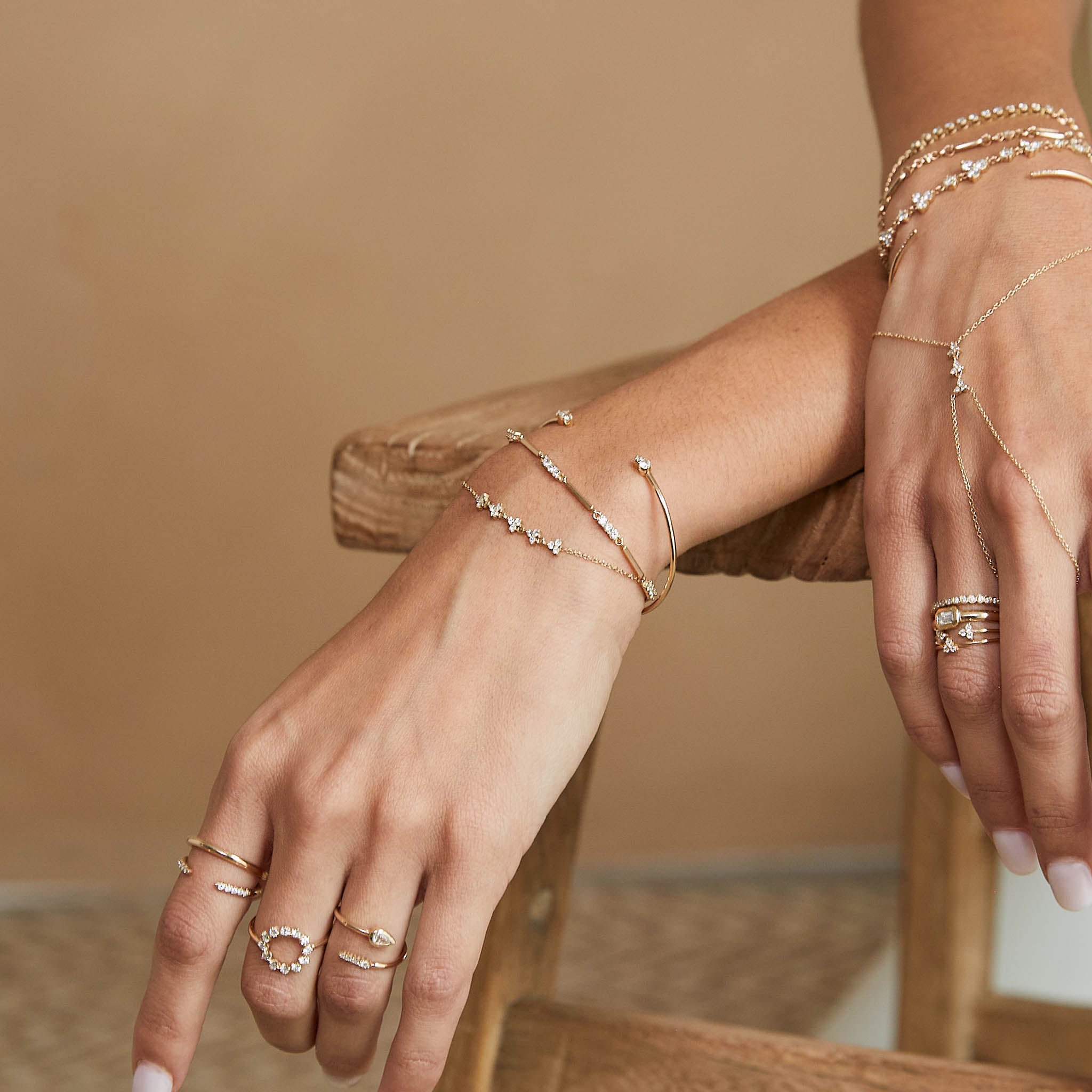 woman with her hands resting on a wooden stool wearing a Zoë Chicco 14k Gold 5 Linked Diamond Trio Bracelet on her wrist  stacked with other bracelets