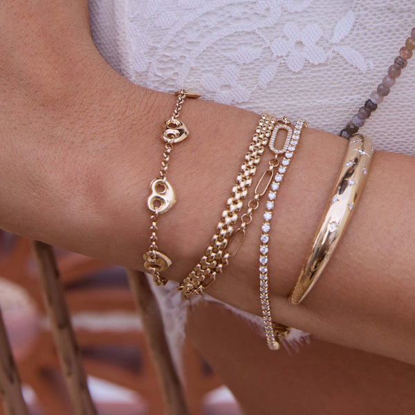 Close-up of a write wearing Zoe Chicco 14k gold and diamond star-set aura cuff, panther chain, and heart marina chain bracelets on a neutral background