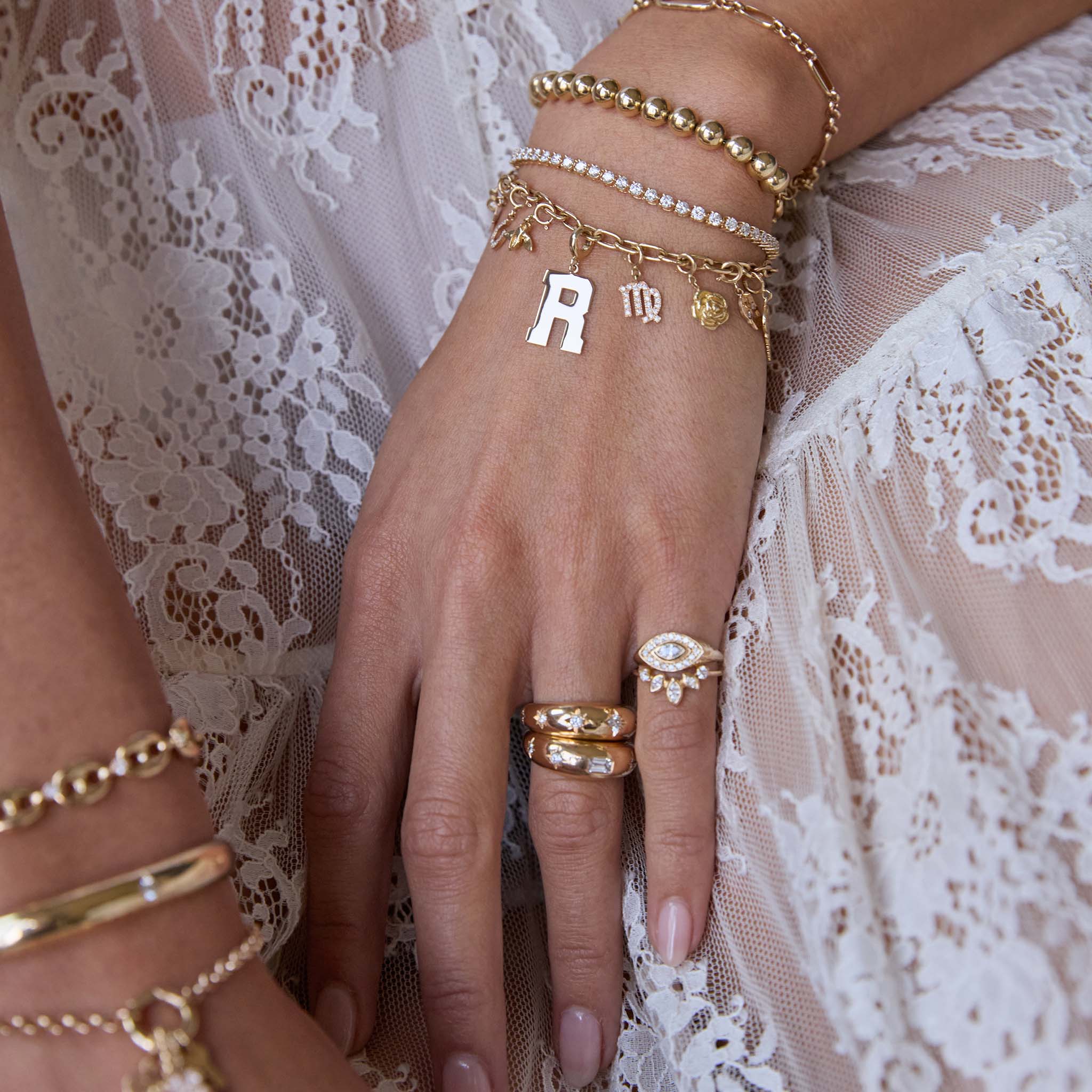 Close-up of a hand wearing multiple Zoe Chicco 14k gold charm bracelets and diamond rings on a white lace background