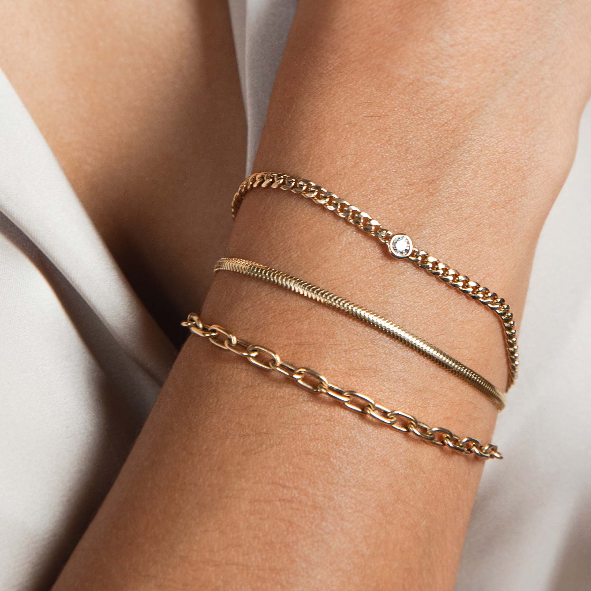 A woman wears 3 bracelets stacked:  a small curb chain with floating diamond, small snake chain and medium square oval chain bracelets.