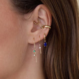 Close-up of an ear with multiple Zoe Chicco 14k gold earrings with green, white, and dark blue gemstones on a neutral background.